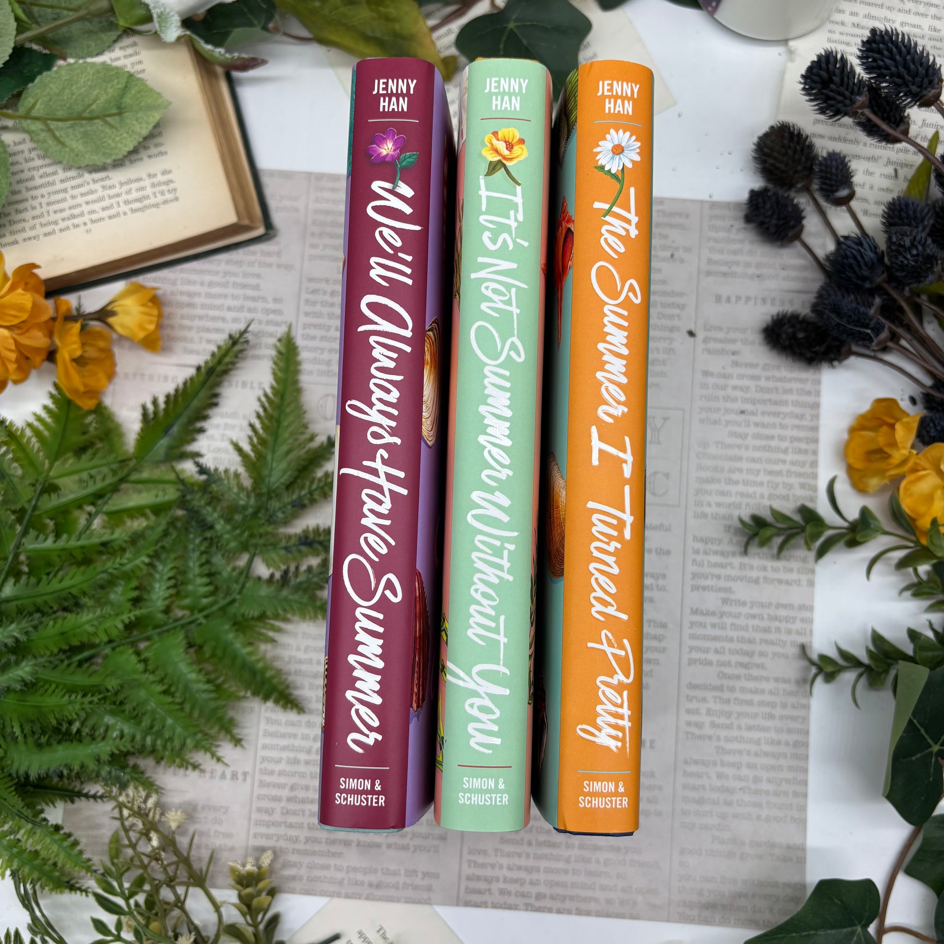 Three books titled 'It's Not Summer Without You', 'The Summer I Turned Pretty', and 'We'll Always Have Summer' by Jenny Han on a textured surface with floral and leaf decorations.