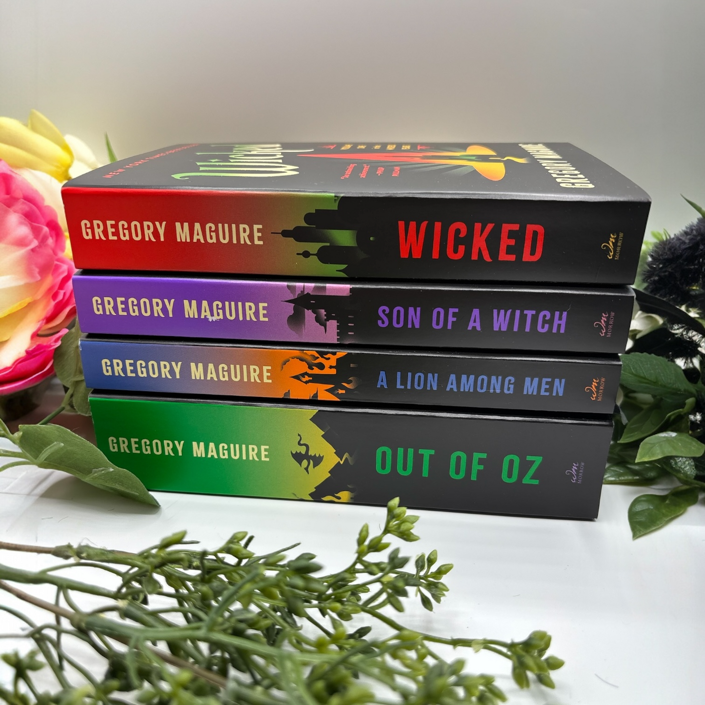 Stack of books by Gregory Maguire with colorful covers on a white surface with flowers and greenery.